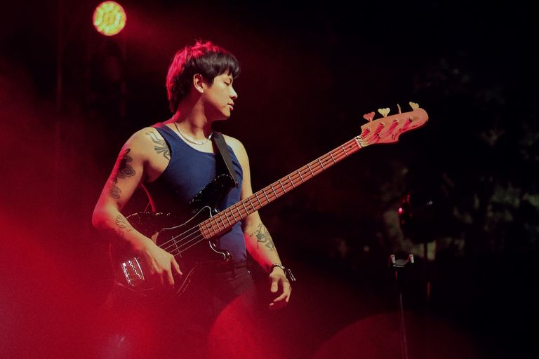 Live performance shot of musician Don Glori playing a bass guitar on stage under warm red concert lighting.